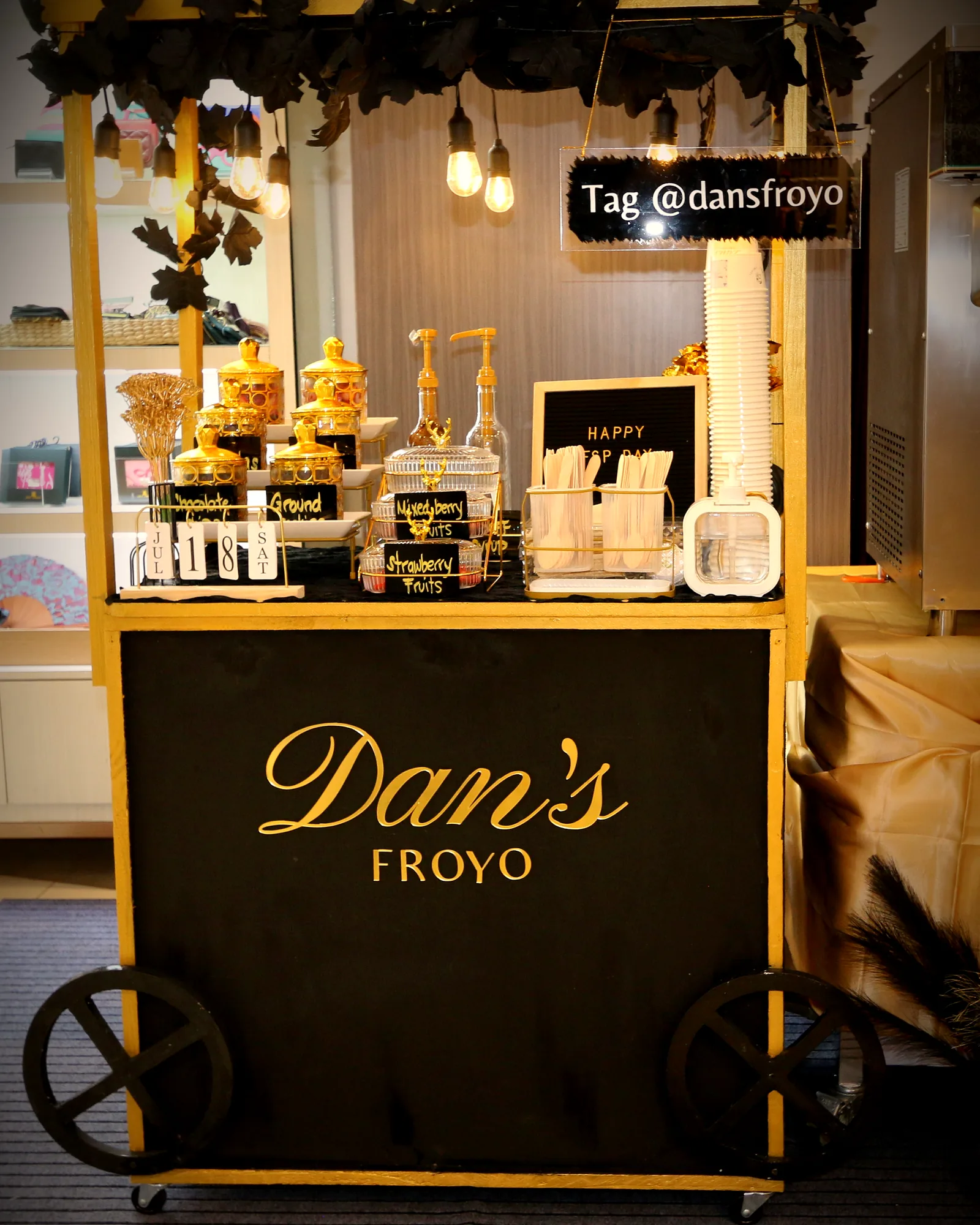 The Luxe cart in matte black with gold script and warm Edison bulbs, styled with a dark foliage garland